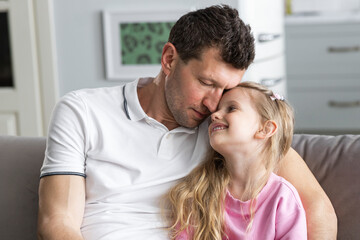 A little daughter sits on the sofa in the room next to her falling asleep father, who hugs her. A...
