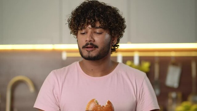 Fast food enjoyment. Young guy eats unhealthy hamburger with french fries at kitchen. Adult man relaxing listen headphones satisfaction american style burger. Person celebrating cheat meal