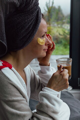 Fototapeta premium Young woman with eye patches and towel drinking morning coffee. Girl after bath with skin mask. Skin care at home. Healthy lifestyle. Pleasure procedure after taking bath. Home cosmetology. 