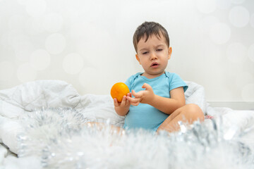Fototapeta na wymiar adorable baby boy on bed blanket with tangerine in hand or candy cane in heart shape. serious funny kid making grimaces,half closed eyes,cool child.merry christmas holiday at home