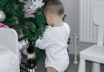 cute baby boy decorating christmas artificial tree inside home house. kid child in white bodysuit...