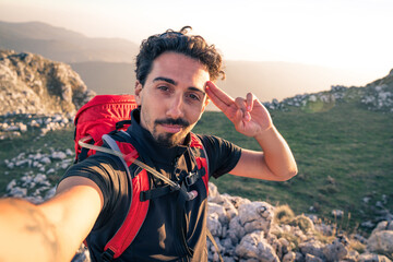 Man takes a selfie in nature path with red backpack exploring new places with amazing sight. He walks on mountains admiring the beauty of nature: rocks and meadows during sunset