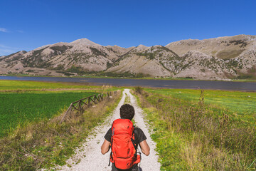 Man walking in nature path with backpack on shoulders exploring new places with amazing sight. He walks through fields along the banks of a river admiring the beauty of nature: meadow and mountains
