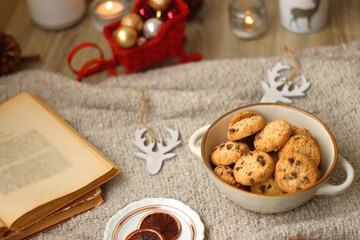 Obraz na płótnie Canvas Bowl of cookies, cup of tea, dry oranges, pine cones, books, reading glasses, small presents, various Christmas decorations and lit candles on the table. Cozy Christmas hygge. Selective focus.