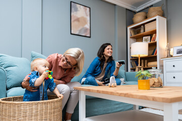 Fototapeta Young irresponsible mother watch TV and eat popcorn while her mother child grandmother looks after the baby. Carefree mom pays no attention to her child who is being looked after by older babysitter obraz