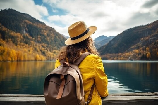 Rear view of a stylish girl, with a backpack, a hat and a yellow jacket, looking at the view of the mountains and the lake in the autumn nature