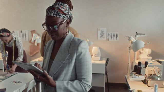 Waist up tracking shot of stylish African American tailoring atelier owner with glasses and tablet walking around controlling work process during workday