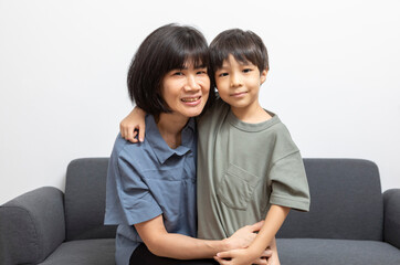 Portrait of happy Asian woman and her little son hugging and sitting on sofa at home.