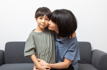 Portrait of happy Asian woman and her little son hugging and sitting on sofa at home.