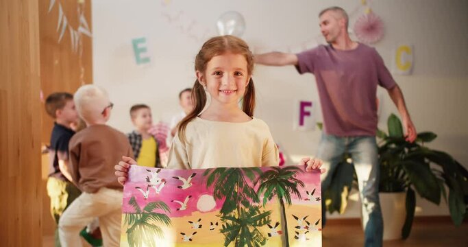 Portrait: a beautiful preschool girl shows her picture that she drew against the backdrop of recess in a club for preschool children to prepare for school. little girl preschool artist shows her