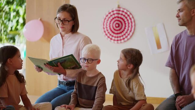 A blonde woman with a boxy hairstyle and a pink shirt reads a green book to preschool children in a special club for preparing children for school