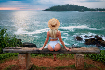 Woman in bikini with a beautiful physique sitting on beach looking at ocean view on sunset in...