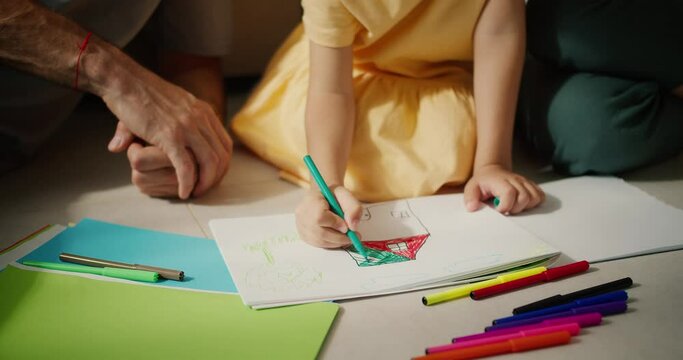 Close-up shot of a little girl in a yellow dress drawing a house using multi-colored markers on a piece of white paper while sitting on the floor with her parents in a modern room
