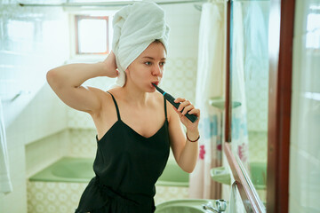 Young adult woman in bathroom with towel on head, promotes dental hygiene using electric toothbrush, mirror reflection shows daily health routine.