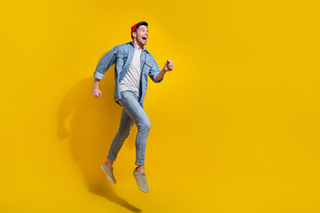 Fototapeta na wymiar Full length photo of cheerful excited guy wear denim jacket running fast jumping high empty space isolated yellow color background