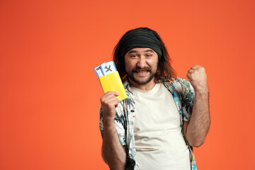 A longhaired man in a bandana holds a passport with airline tickets and makes a victory gesture with his hand. Happy man in studio on orange background close up. - obrazy, fototapety, plakaty