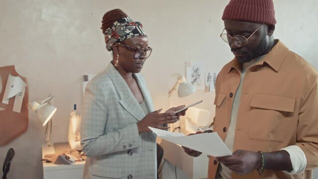 Medium shot of Black male fashion designer speaking and showing sketches to African American female atelier owner in glasses who then going away