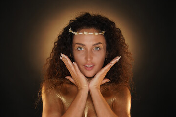 Portrait of female model in studio. Attractive girl wearing tiara and bright makeup covered in golden paint holding face in hands smiling at camera.