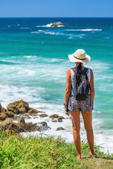 Young woman in a hat wearing sports clothes with a backpack stands on a cliff, looks down at the blue ocean and rocks at the shore, seaside landscape on a summer sunny day.