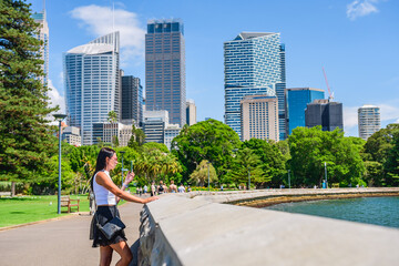 A young woman in a T-shirt and skirt stands in the park on the promenade and talks on the phone, skyscrapers in the business district, view from the Royal Botanic Garden Sydney, summer sunny day. - obrazy, fototapety, plakaty