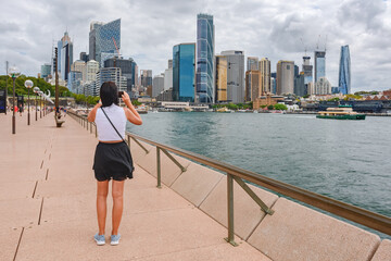 Young woman in a T-shirt and skirt takes a photo with her phone of Circular Quay, skyscrapers in the business district, view from the Sydney Opera House, summer cloudy day.
