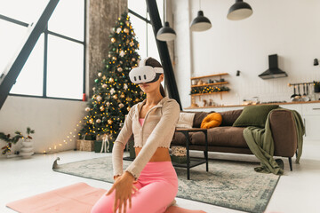 A young woman in pink sportswear practices yoga in a virtual reality headset against the backdrop of a Christmas tree.