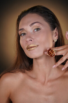 Portrait of brunette female model in studio. Attractive woman with hairstyle and makeup posing with golden rose flower on face looking at the camera.