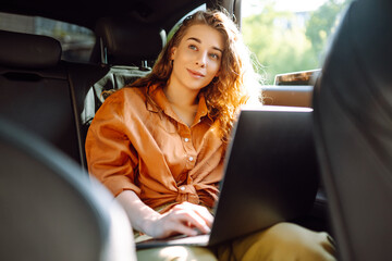 Beautiful business woman is using a laptop while sitting in the back seat of a car. Young freelancer woman working on a laptop in a car. Freelance, transportation concept.