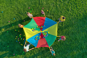 Group of children and teachers playing with rainbow playground parachute on green grass, top view....