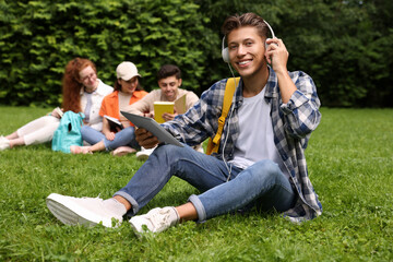 Students learning together in park. Happy young man with headphones working with tablet on green grass, selective focus