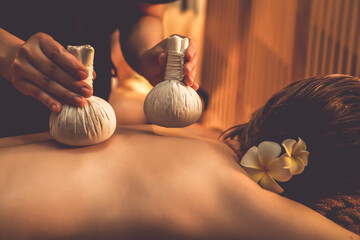 Hot herbal ball spa massage body treatment, masseur gently compresses herb bag on couple customer body. Serenity of aromatherapy recreation in warm lighting of candles at spa salon. Quiescent - obrazy, fototapety, plakaty