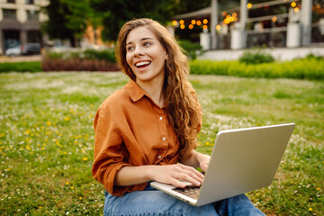 Curly-haired woman in casual clothes with a laptop sits on the lawn on a sunny day. Happy freelancer woman working outdoors and enjoying the weather.