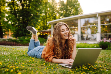 Curly-haired woman in casual clothes with a laptop sits on the lawn on a sunny day. Happy...