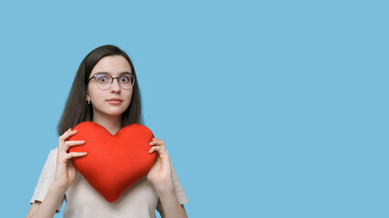Portrait of a beautiful smiling student girl with glasses holding a bright red heart-shaped pillow