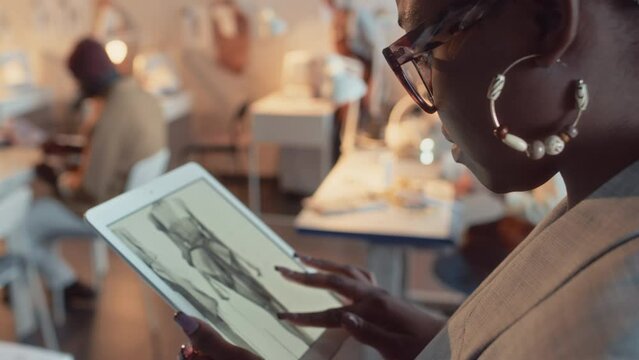 Side closeup of African American female fashion designer looking at sketches of women and men apparel on tablet in tailoring studio