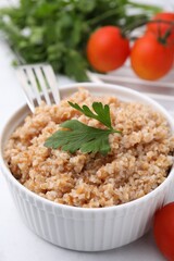 Tasty wheat porridge with parsley and tomatoes in bowl on white table, closeup