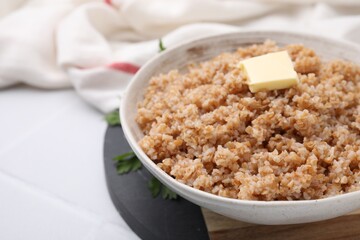Tasty wheat porridge with butter and parsley in bowl on white tiled table, closeup. Space for text