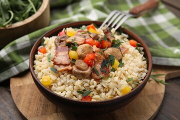 Cooked bulgur with vegetables, fried bacon and mushrooms in bowl on wooden table, closeup