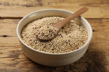 Raw quinoa seeds and spoon in bowl on wooden table, closeup