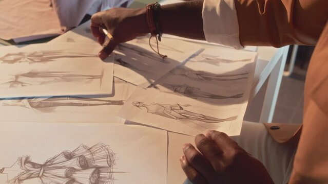 Side close up of unrecognizable clothes designer drawing sketches of new fashion apparel collection sitting at table in atelier
