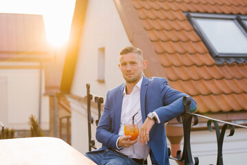 Young handsome man is sitting in an outdoor summer cafe in a blue suit and holding a cocktail glass...