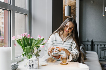 Happy young woman pours tea from a kettle into a mug at a table in a cafe