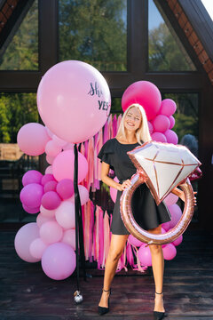 Vertical portrait of elegant blonde female in summer dress posing holding various colorful festive foil helium balloons standing on background of country house building, smiling looking at camera.