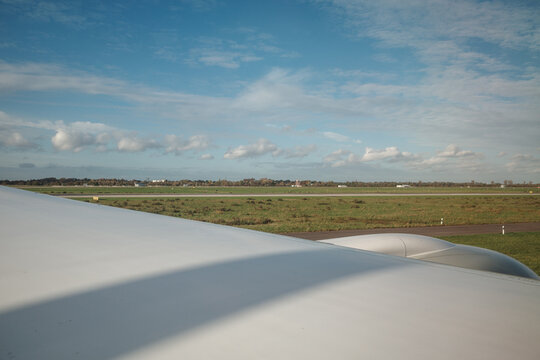 A close-up photograph from inside an airplane, showing the aircraft wing and engine cowl extending over a view of green fields, an airfield, and a blue sky with scattered white clouds during the day.