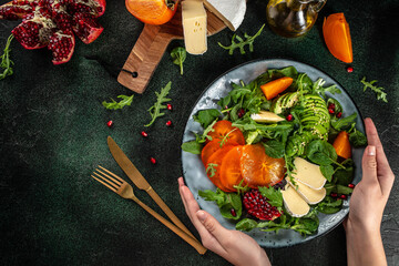 Woman holding Persimmon salad with fresh greens, avocado, Camembert cheese and pomegranate, top...