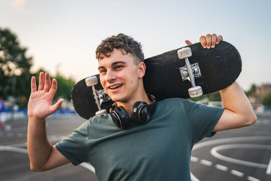 One man young adult caucasian teenager stand outdoor with skateboard on his shoulder and headphones posing at basketball court portrait happy confident wear shirt casual real person copy space