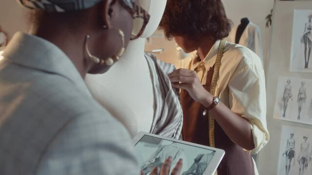 Over shoulder tilt shot of African American creative director and tailor working on new garment looking at sketch on tablet and pinning striped textile to mannequin at sewing workshop