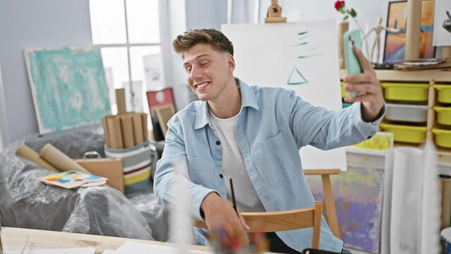 Handsome young caucasian man, confident artist sitting in art studio, making a selfie with smartphone amid his creative painting journey.