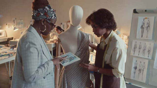 Medium shot of African American female creative director with tablet controlling process of creating dress and young Black tailor pinning striped fabrics to mannequin in atelier