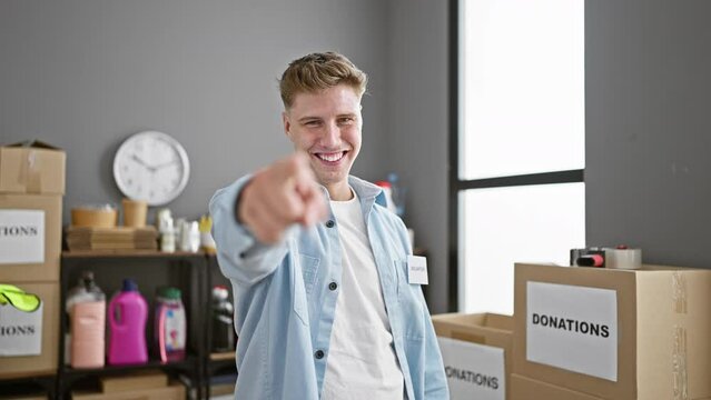 Confident young caucasian man with a charming smile, volunteering at a charity center, enthusiastically pointing to the camera while standing amidst donation boxes